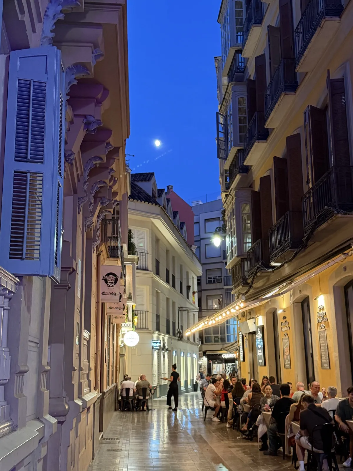 A narrow street in Málaga's old town at night, lit by street lamps, with the moon overhead and people eating at outdoor tables