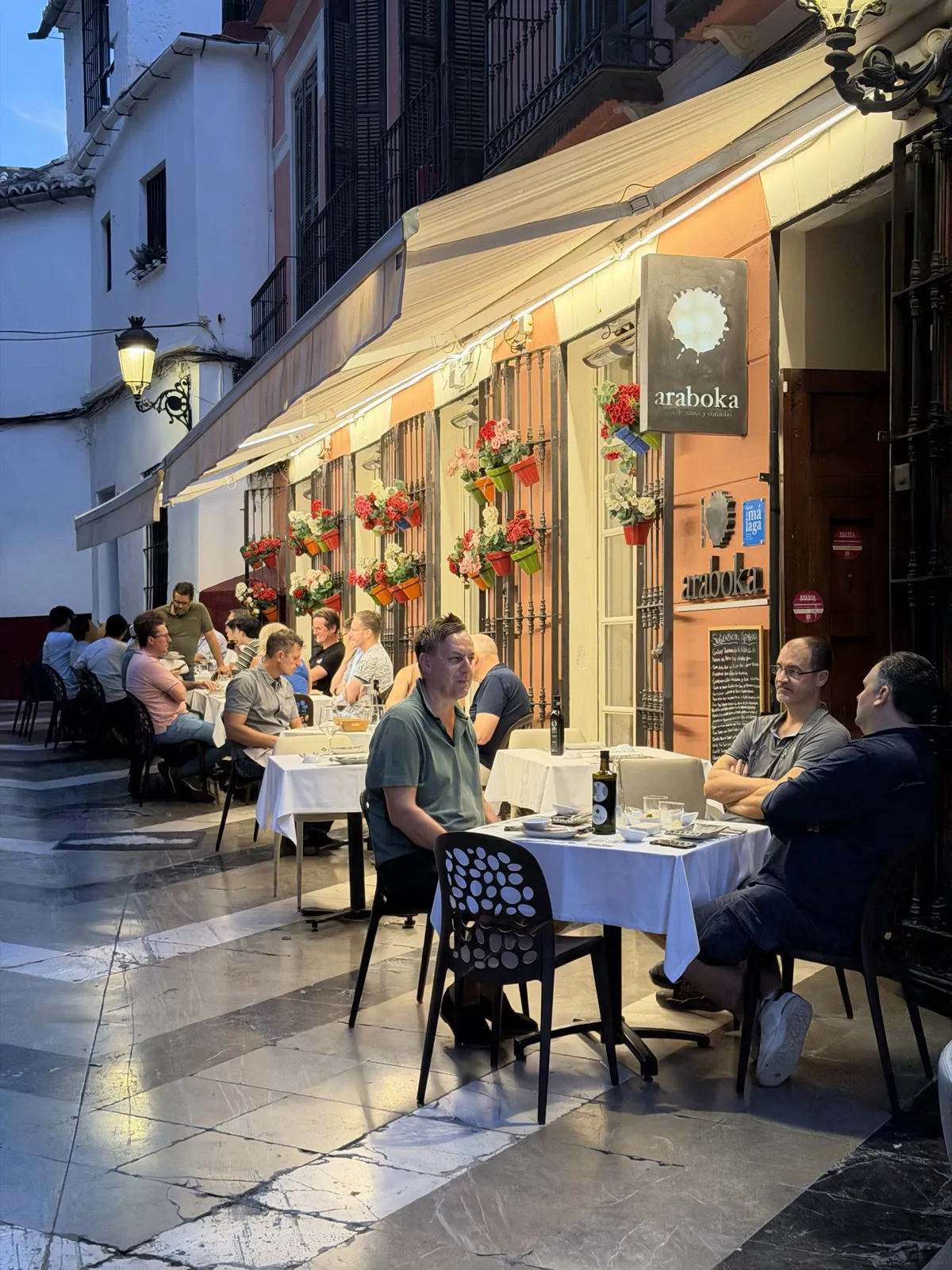 Diners at white-clothed tables on a restaurant terrace in Málaga's old town at dusk, with flower boxes on the wall behind
