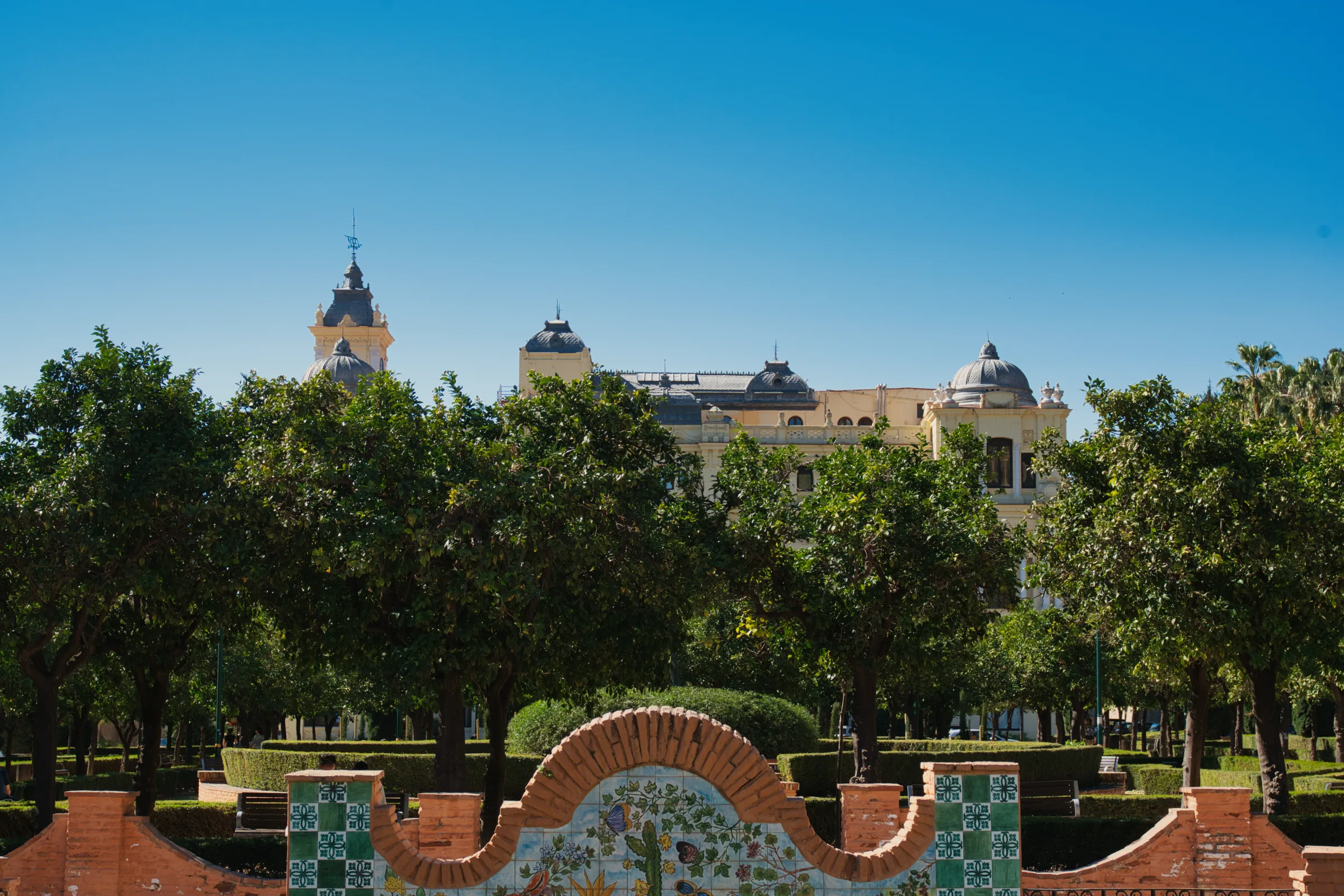 Malaga's Ayuntamiento building seen through the trees of Parque de Málaga, with a tiled bench in the foreground