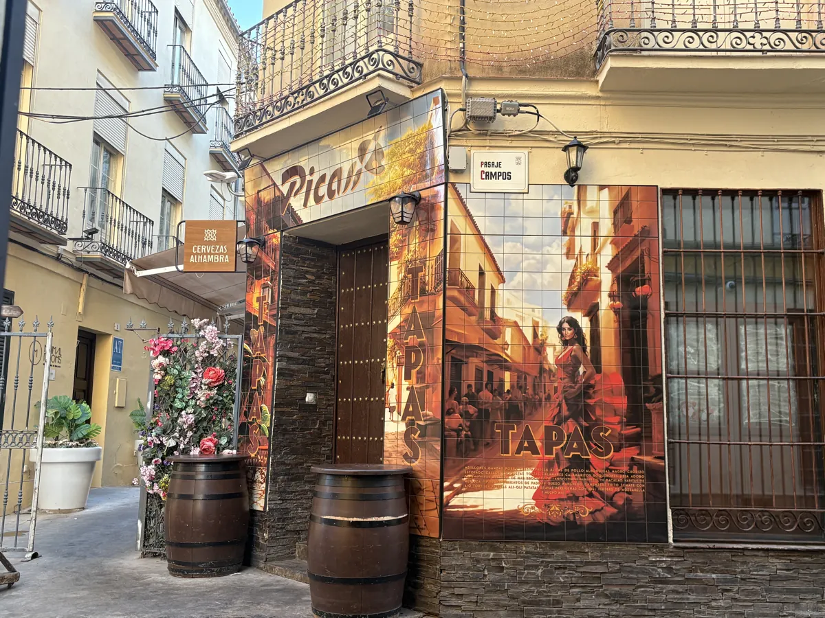 Picas Bar's tiled corner facade on Pasaje Campos, hand-painted with a flamenco dancer and a Málaga street scene, TAPAS lettered down the tiles above wine barrels used as tables
