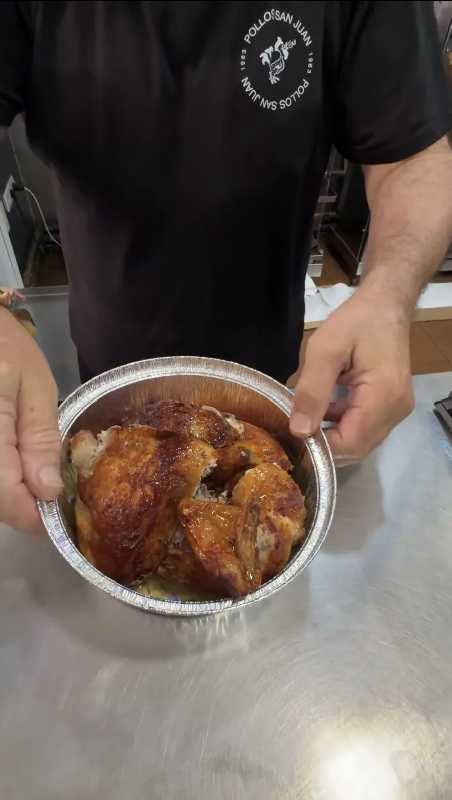 A staff member holding a foil tray of whole roast chicken at Pollos San Juan in Málaga