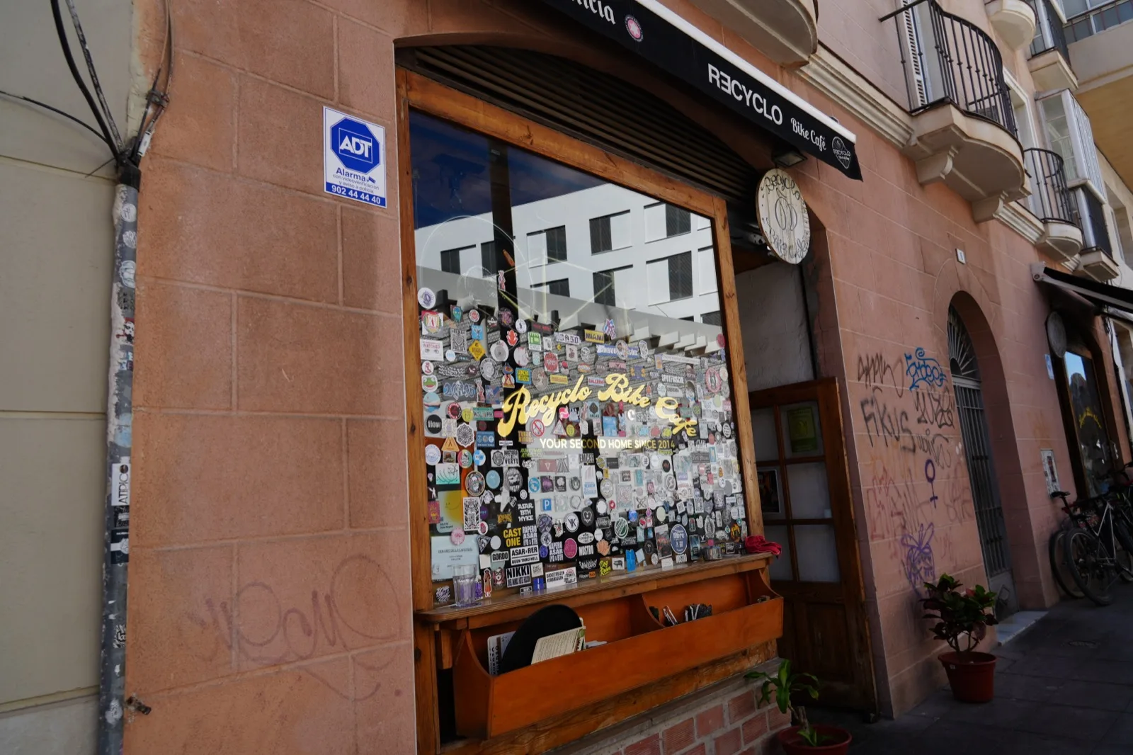 The sticker-covered front window of Recyclo Bike Café in Málaga, with "your second home since 2014" painted in gold lettering