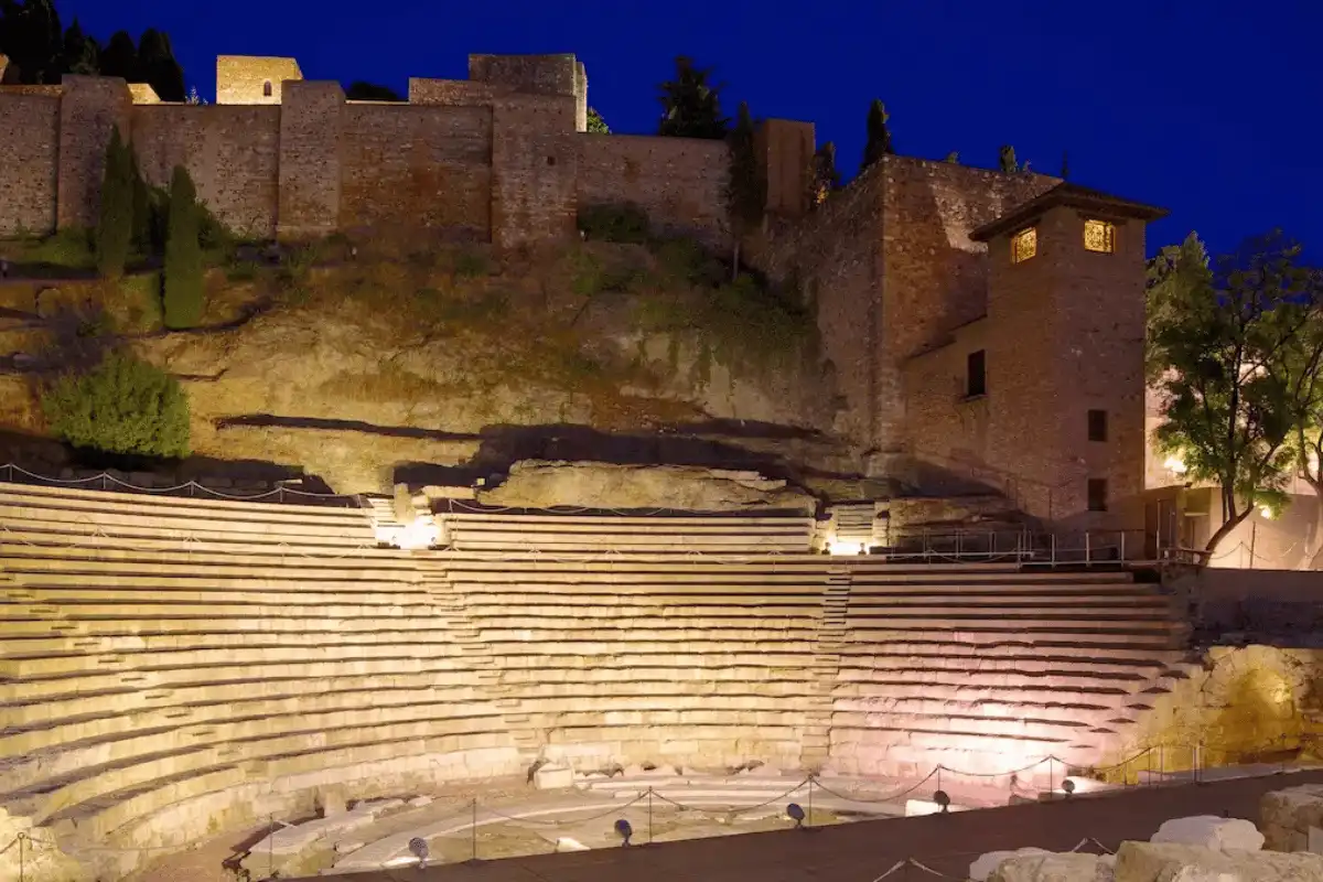 The stone seating of Málaga's Roman Theatre lit up in the evening, with the Alcazaba walls above it