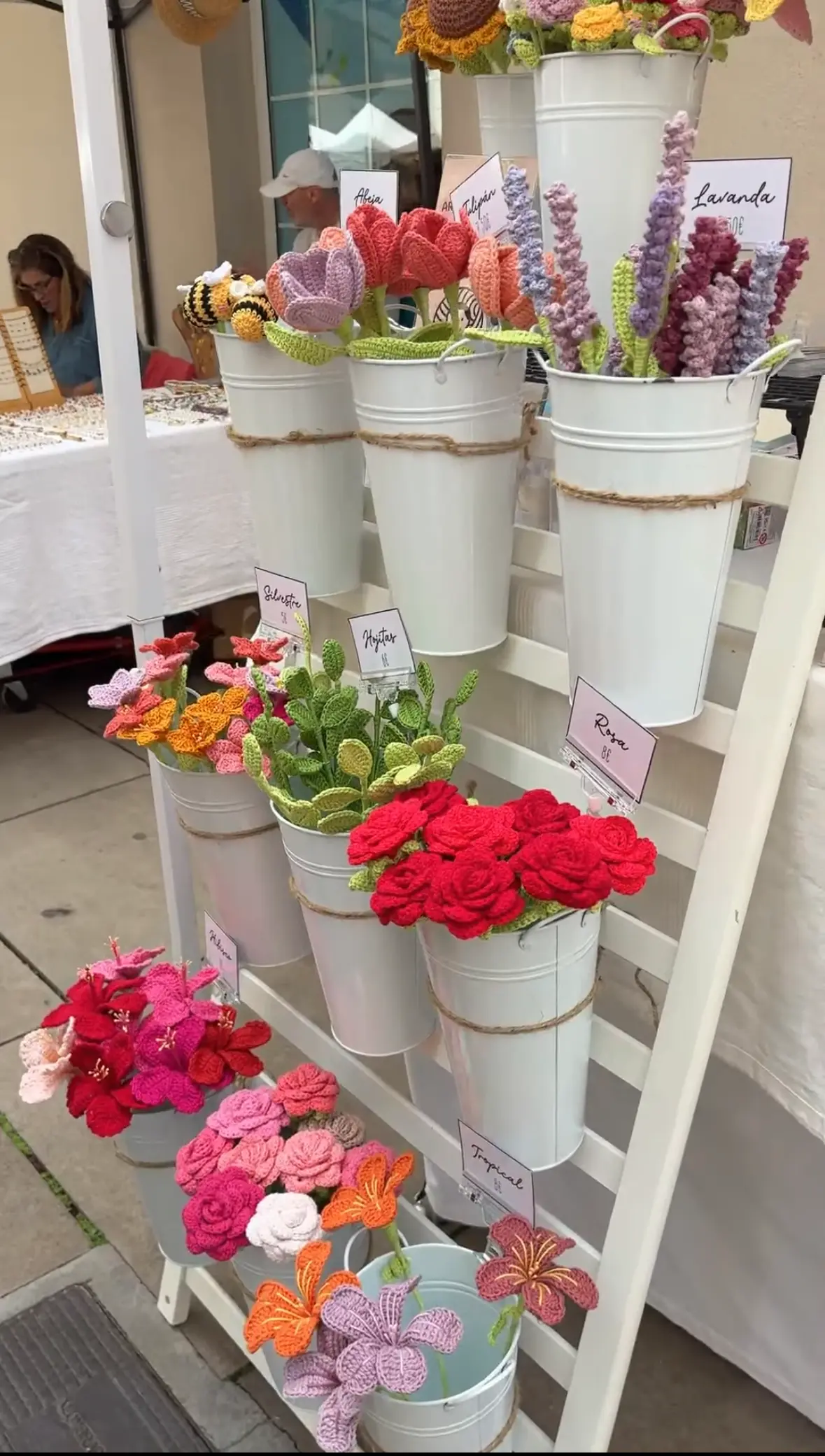 Rows of handmade crochet flowers in white buckets, labelled with names like tulipán, lavanda, and rosa, at the Pom Pom stall in Malaga's Soho market.