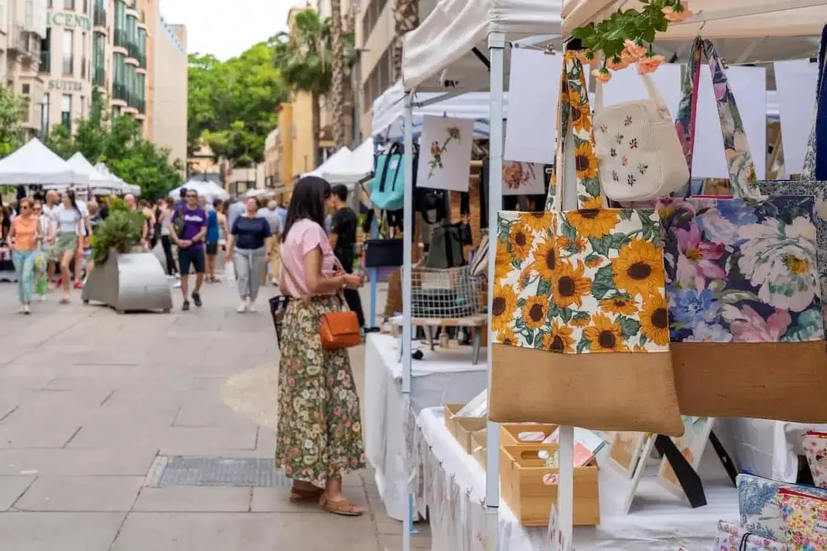 A shopper browsing market stalls hung with floral tote bags and prints along a tree-lined street at Rastro Cultural Soho in Malaga