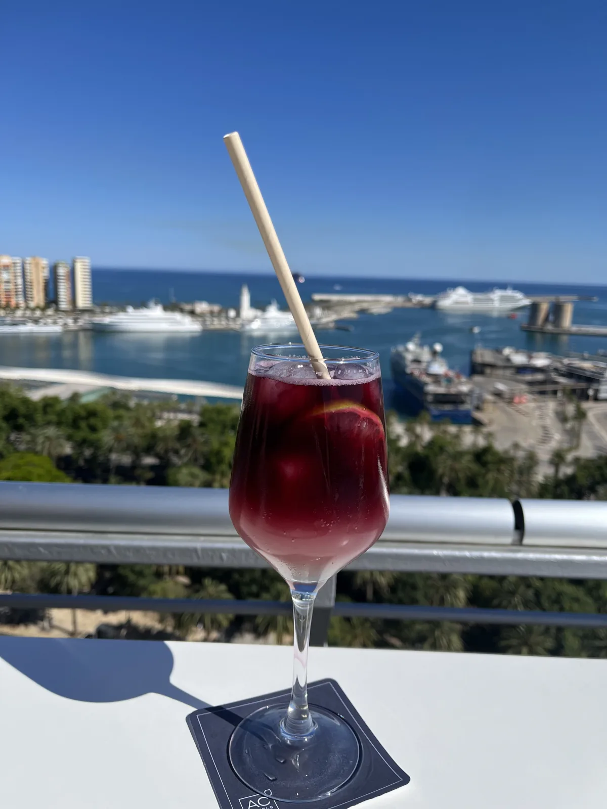 A glass of tinto de verano with a paper straw and a slice of lemon on a rooftop bar table, the port of Málaga and its boats below