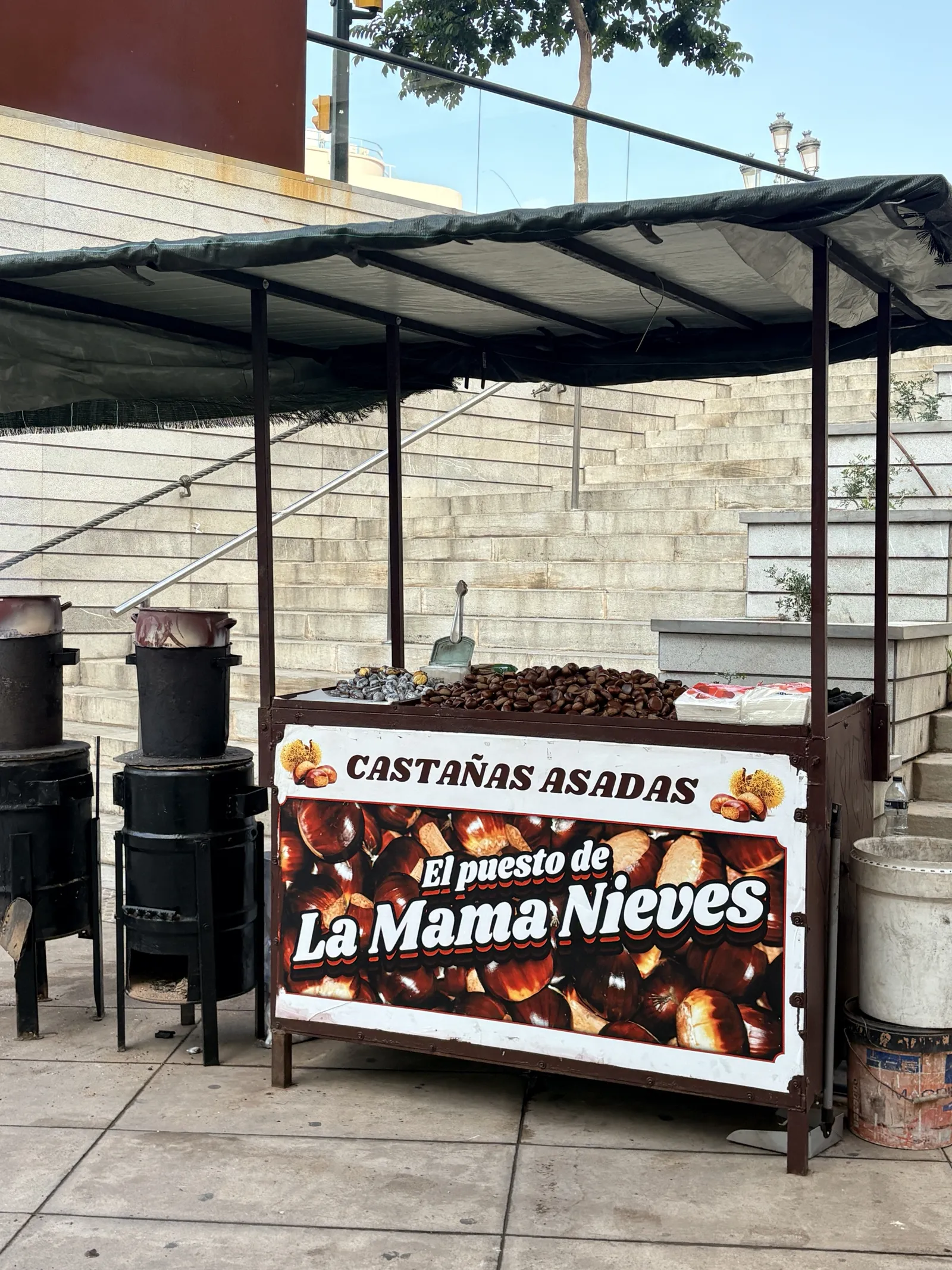Castañas asadas stand El puesto de La Mama Nieves at the foot of the Tribuna de los Pobres steps in Málaga, with chestnuts piled on the cart under a green canopy and two black chestnut-roasting drums beside it
