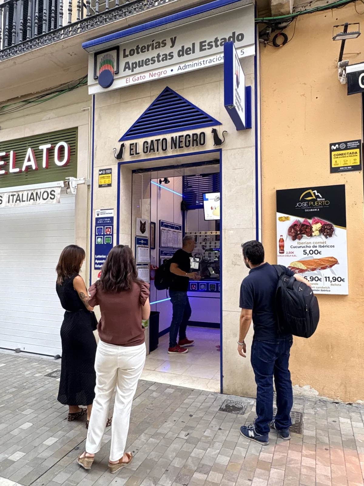 Entrance to El Gato Negro state lottery shop on Calle Granada in Málaga, with black cat signs above the door and three people standing outside looking in