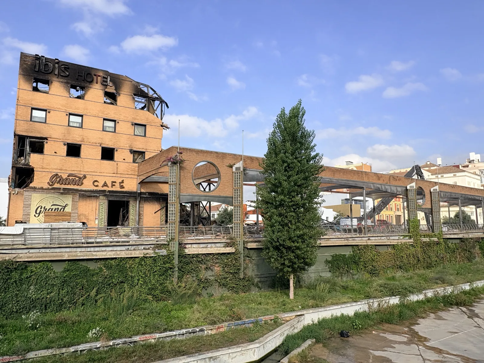 Fire-damaged Ibis Hotel and Grand Café on Pasillo de Guimbarda in Málaga, seen from the Guadalmedina riverbed, with blackened upper floors and demolition machinery working behind
