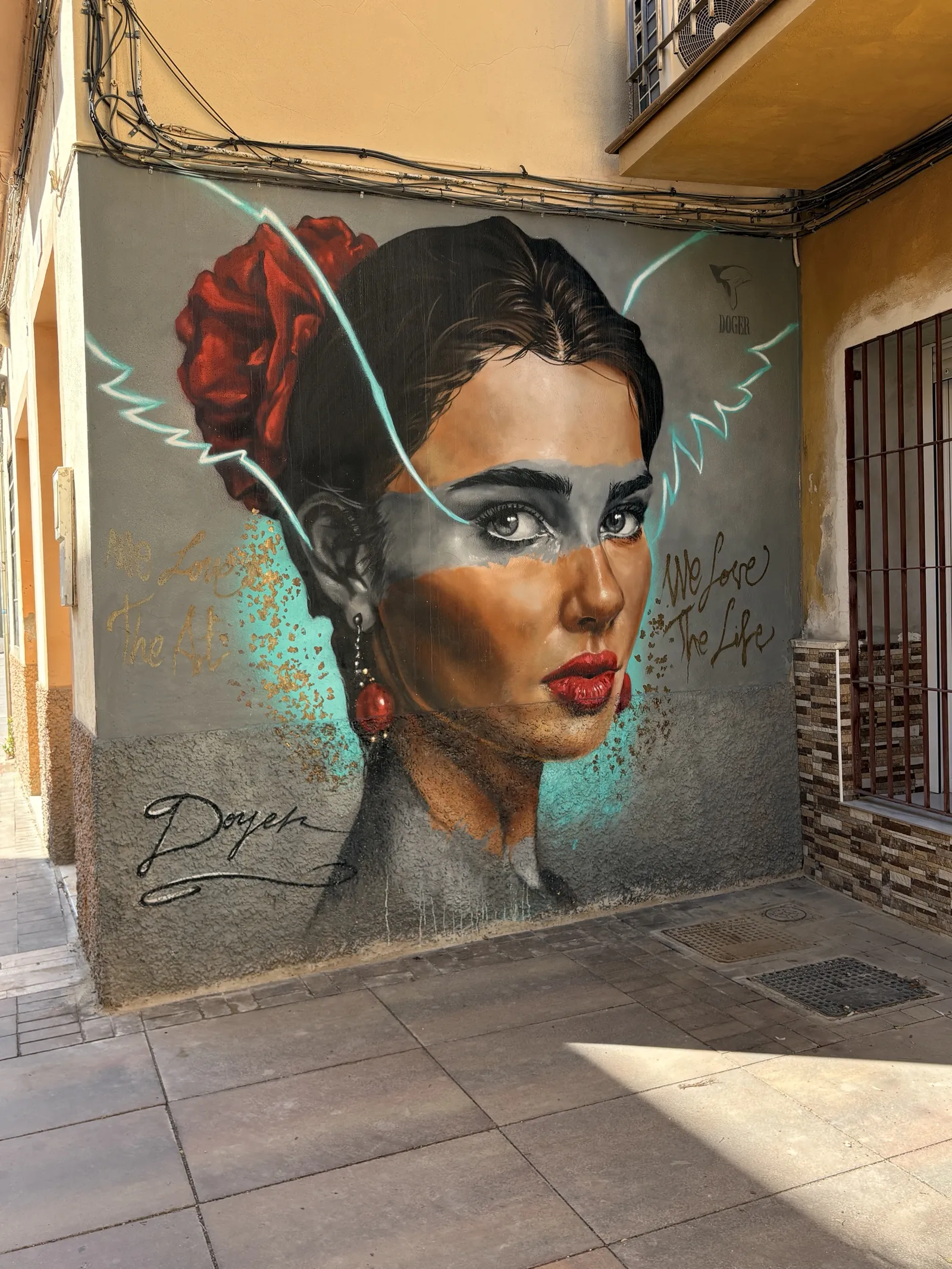 Street mural by Doger in the Lagunillas neighbourhood of Málaga: a large portrait of a woman with a red rose in her hair, red earrings and red lips, a grey band painted across her eyes, turquoise wing-like lines and the gold words We Love The Art and We Love The Life, on a grey corner wall under tangled cables and a balcony