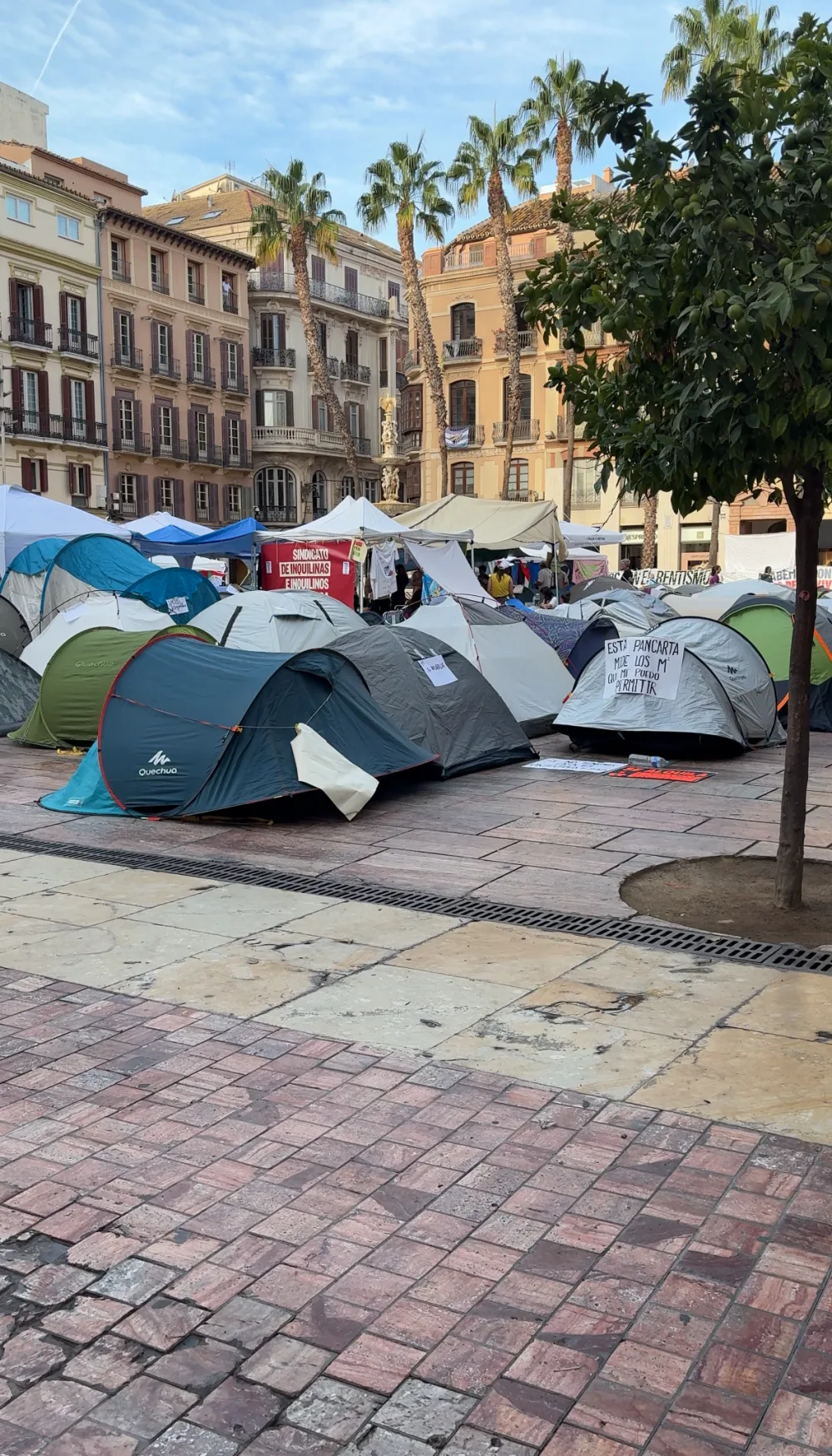 Camping tents filling Plaza de la Constitución in Málaga during a housing protest, with a red tenants' union banner and palm trees behind