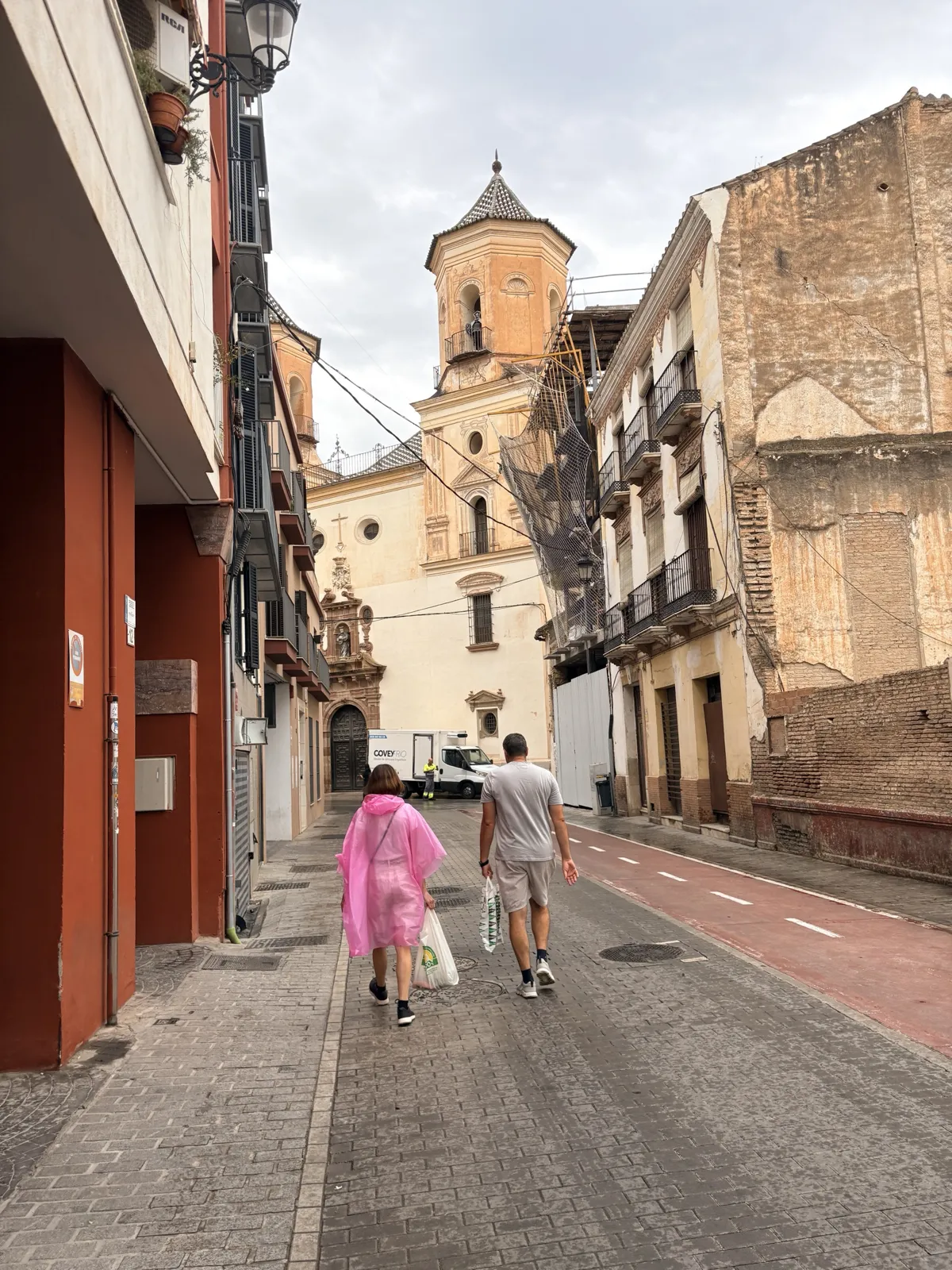 Two people walking along a wet cobbled street in Málaga's old town after rain, one in a pink rain poncho, towards the orange octagonal bell tower and cream façade of the church of San Felipe Neri