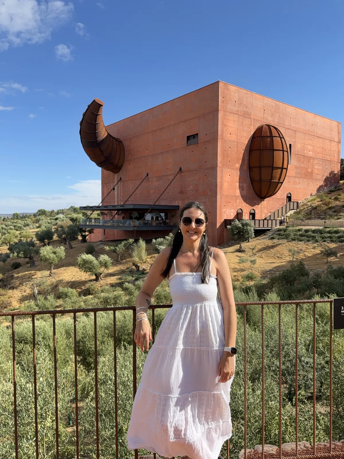 Heidi in a white sundress at the rusty railing of the mirador at LA Almazara near Ronda, with the red cube-shaped olive oil mill behind her, its steel bull's horn, oval window and chain-hung terrace above the olive groves