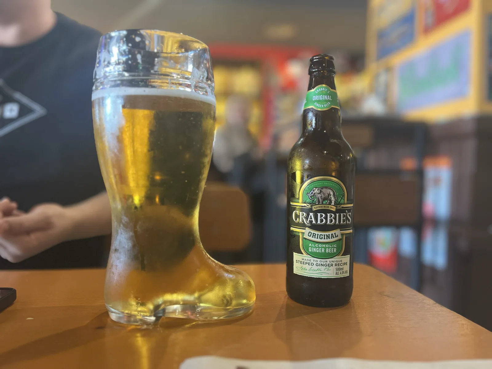 A glass beer boot half full of lager on a wooden table inside Cervecería Untertürkheim, with a bottle of Crabbie's ginger beer beside it