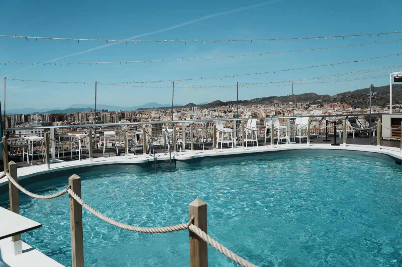The rooftop pool at the AC Hotel Málaga Palacio in full midday sun, string lights overhead and the city and mountains behind