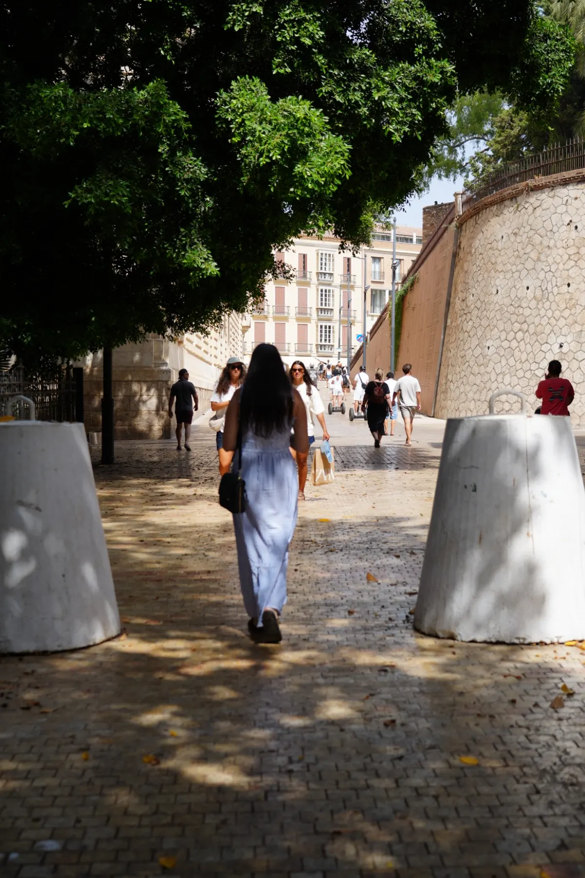 Heidi, a woman with long dark hair in a blue striped sundress, walking up the cobbled approach toward the Alcazaba between white bollards