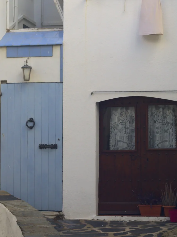 A pale blue plank door with an iron ring knocker next to a dark wooden double door with lace curtains, on a white-walled corner in Cadaqués