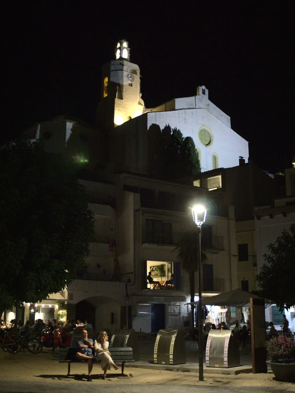 The floodlit white church tower of Cadaqués above the old town at night, a couple sitting on a bench under a street lamp in the square below