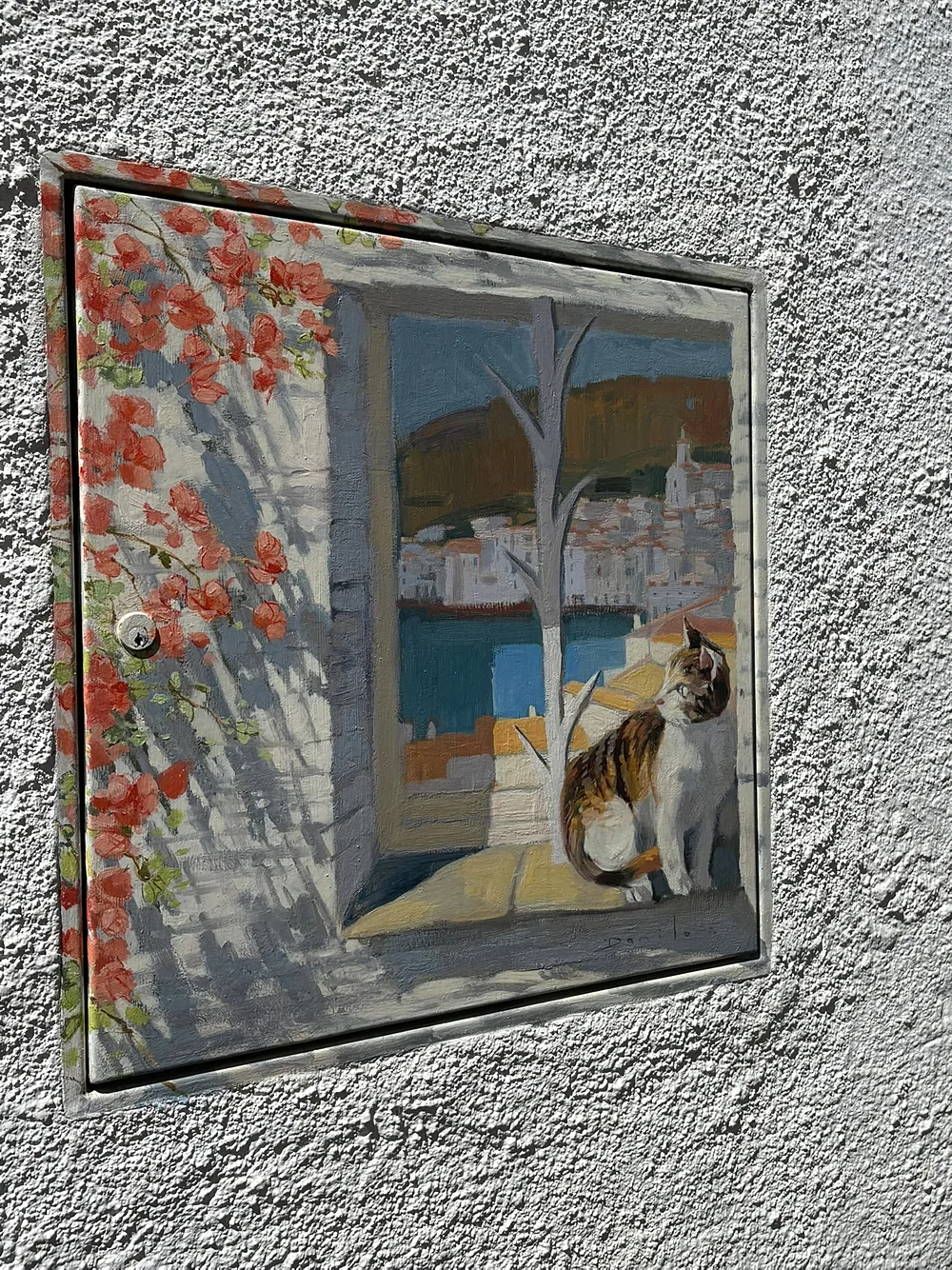 A utility meter cover on a white wall in Cadaqués painted with a cat sitting on a sunny windowsill looking out at the village and bay, red flowers framing the left side