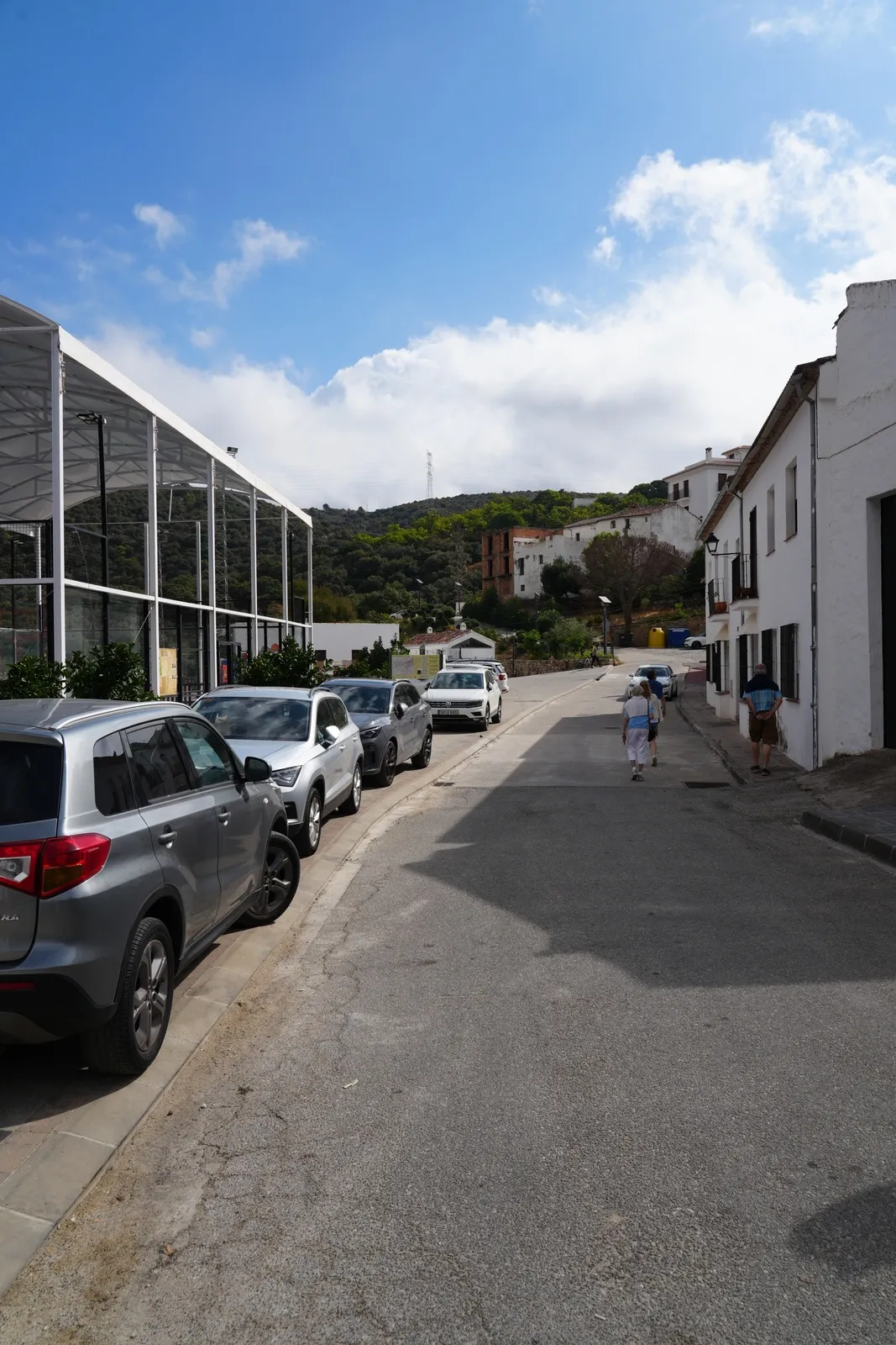 Cars parked along the left side of a narrow village road in Parauta, with white houses on the right and a greenhouse frame at the end of the street