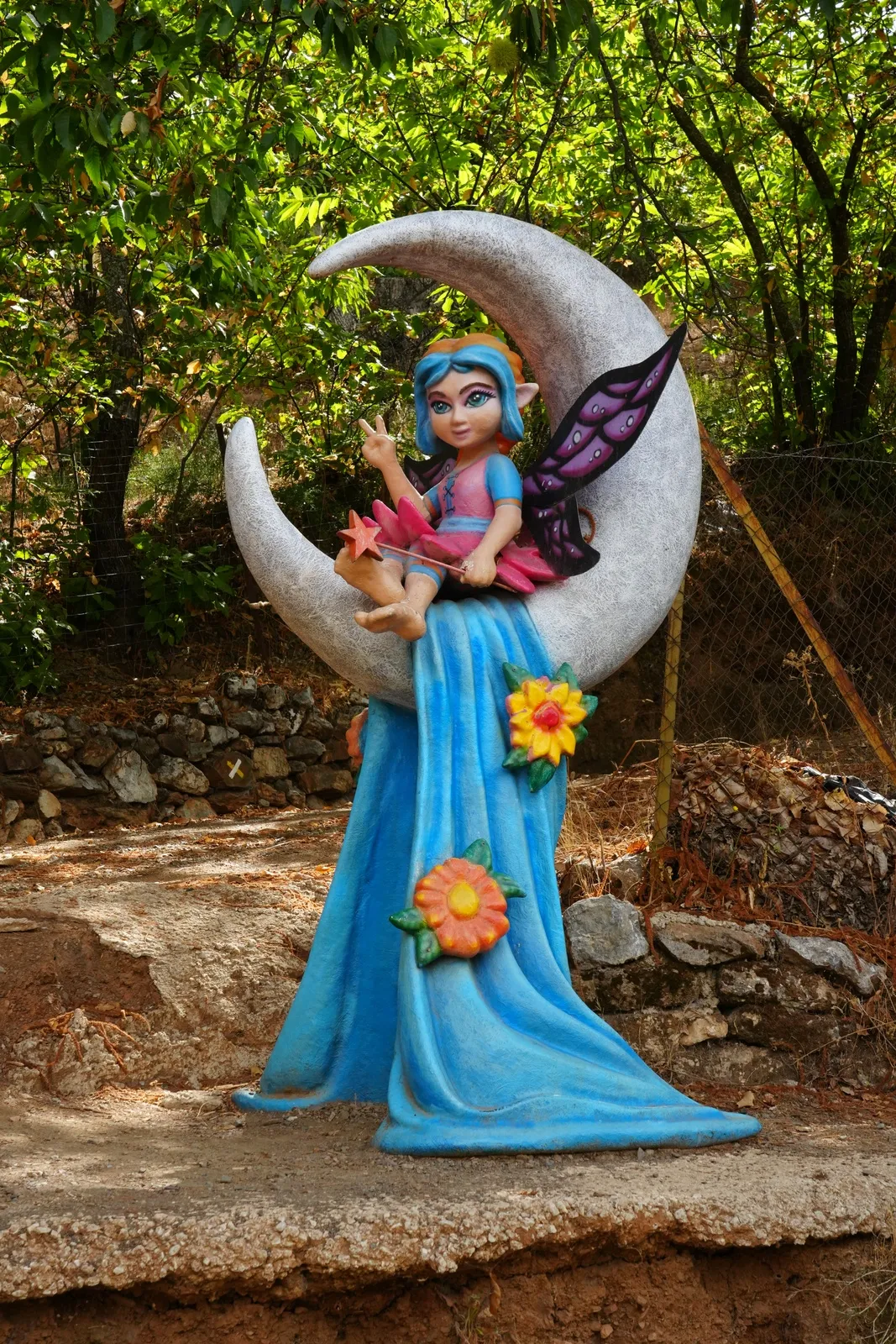 A painted fairy with blue hair and purple wings sitting on a crescent moon, wearing a long blue dress with orange and yellow flowers, beside a dry-stone wall