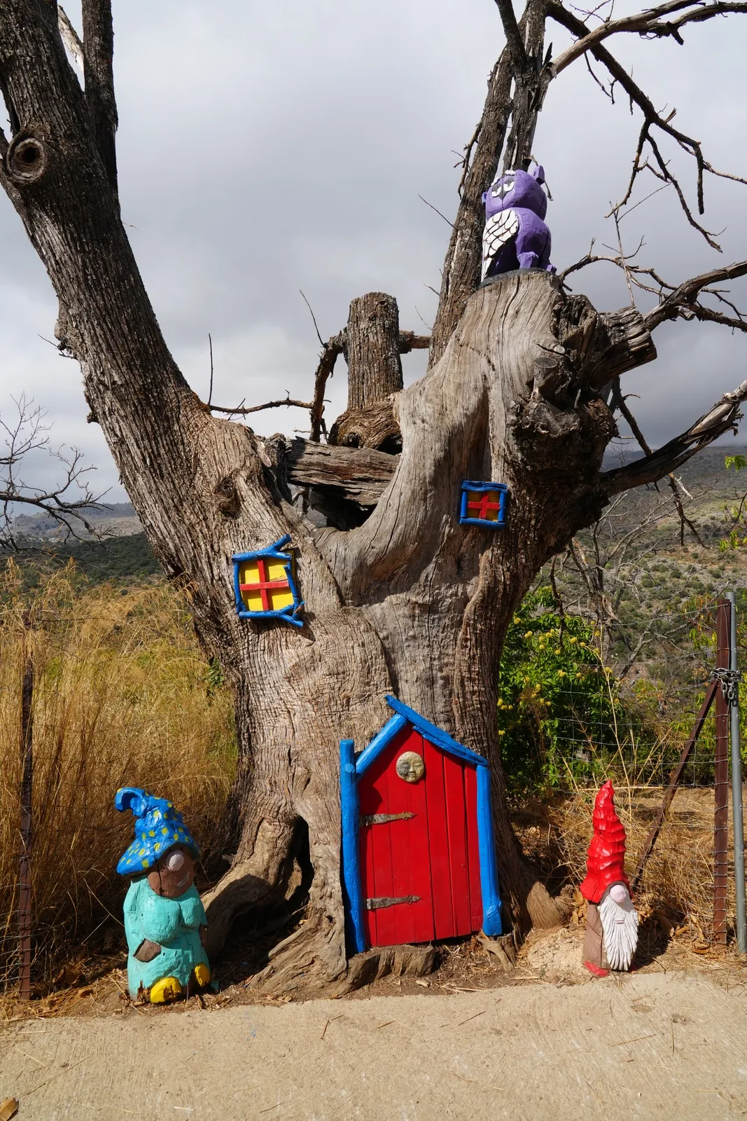 A hollow chestnut trunk with a small red door, tiny windows and a purple owl on top, with a blue-hatted gnome and a red-hatted gnome standing at its base