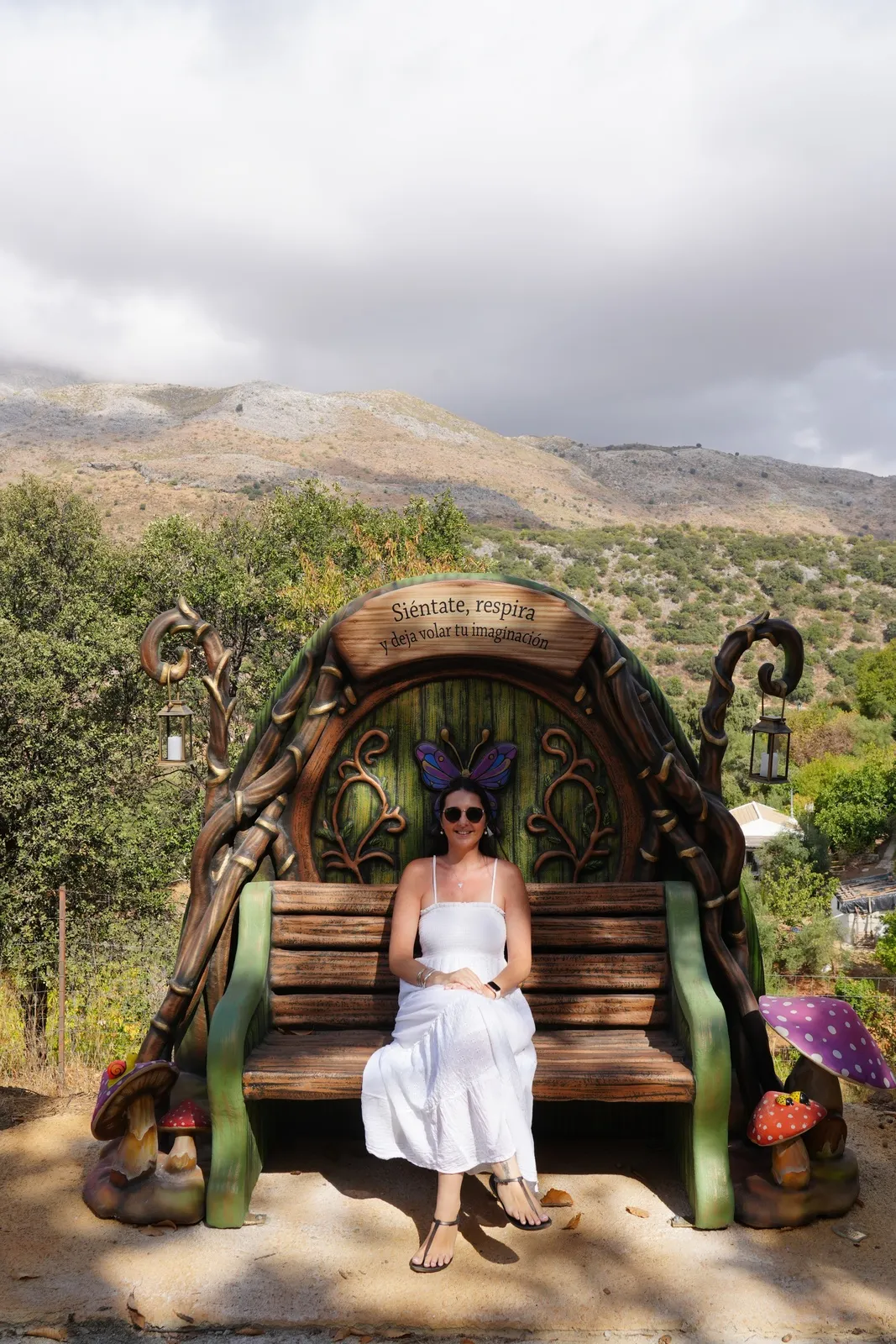 Heidi in a white sundress sitting on a wooden bench under a carved arch with butterfly wings above her and a sign that reads Siéntate, respira, with mountains behind
