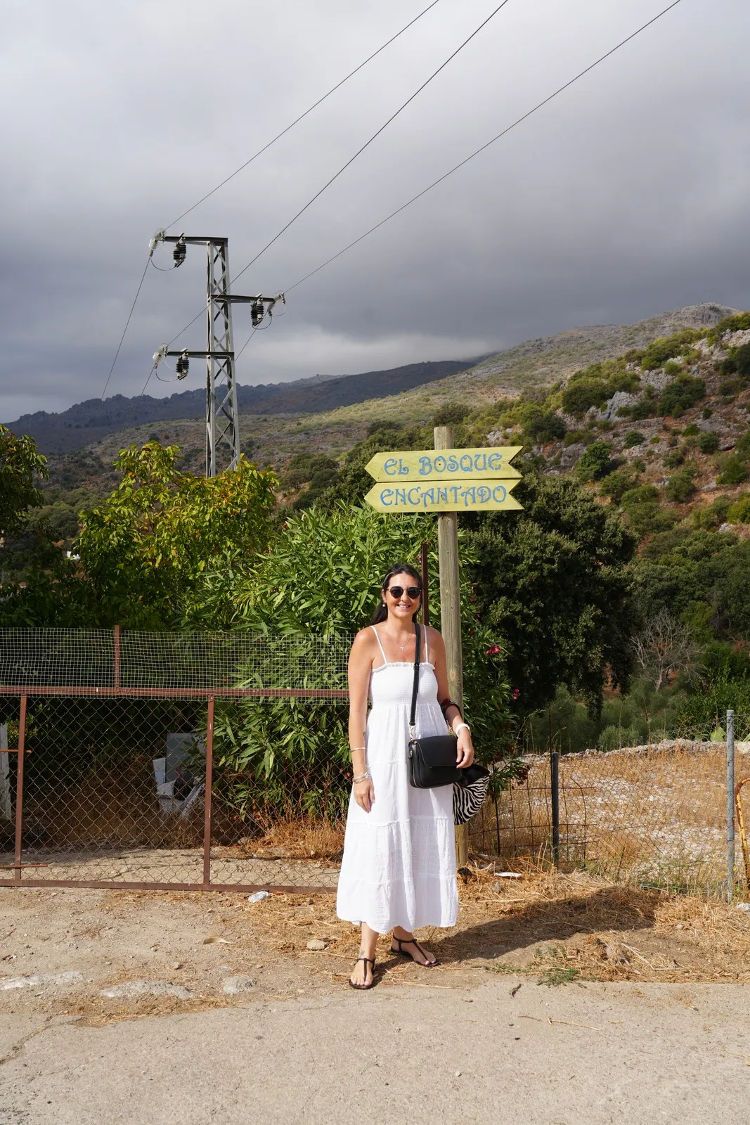 Heidi in a white sundress standing beside a wooden signpost pointing to El Bosque Encantado, with mountains and power lines behind