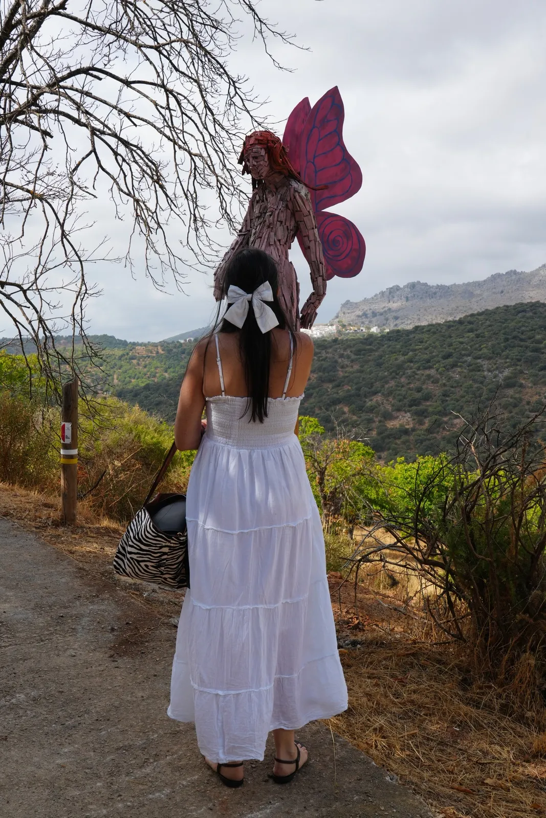 Heidi seen from behind in a white sundress, with a wooden fairy statue with pink wings standing on a post above her head and mountains behind