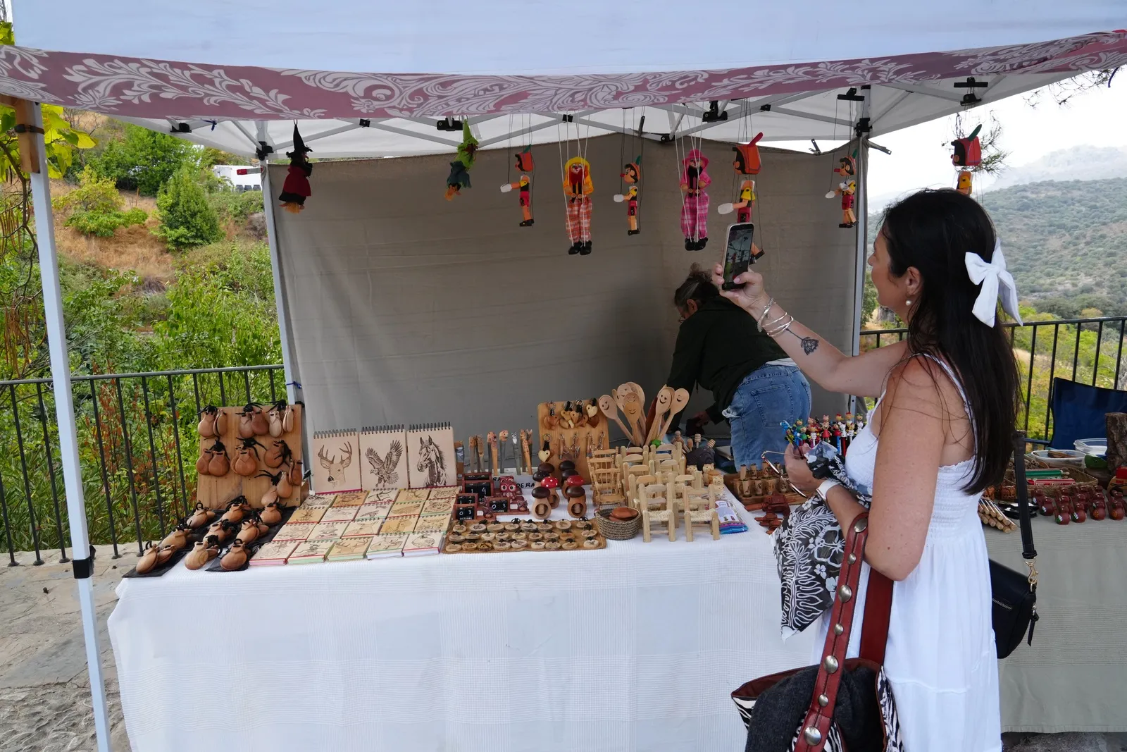 Heidi in a white sundress with a white bow, holding up her phone at a market stall of handmade wooden gifts and puppets hanging under a white canopy
