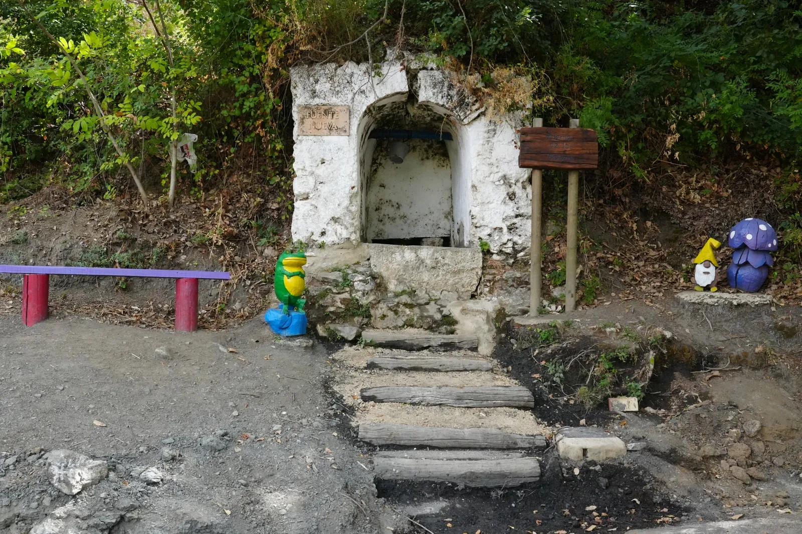 A small whitewashed stone fountain in an alcove with a green frog statue beside the steps, a colourful bench and a wooden sign, in dry earth under trees
