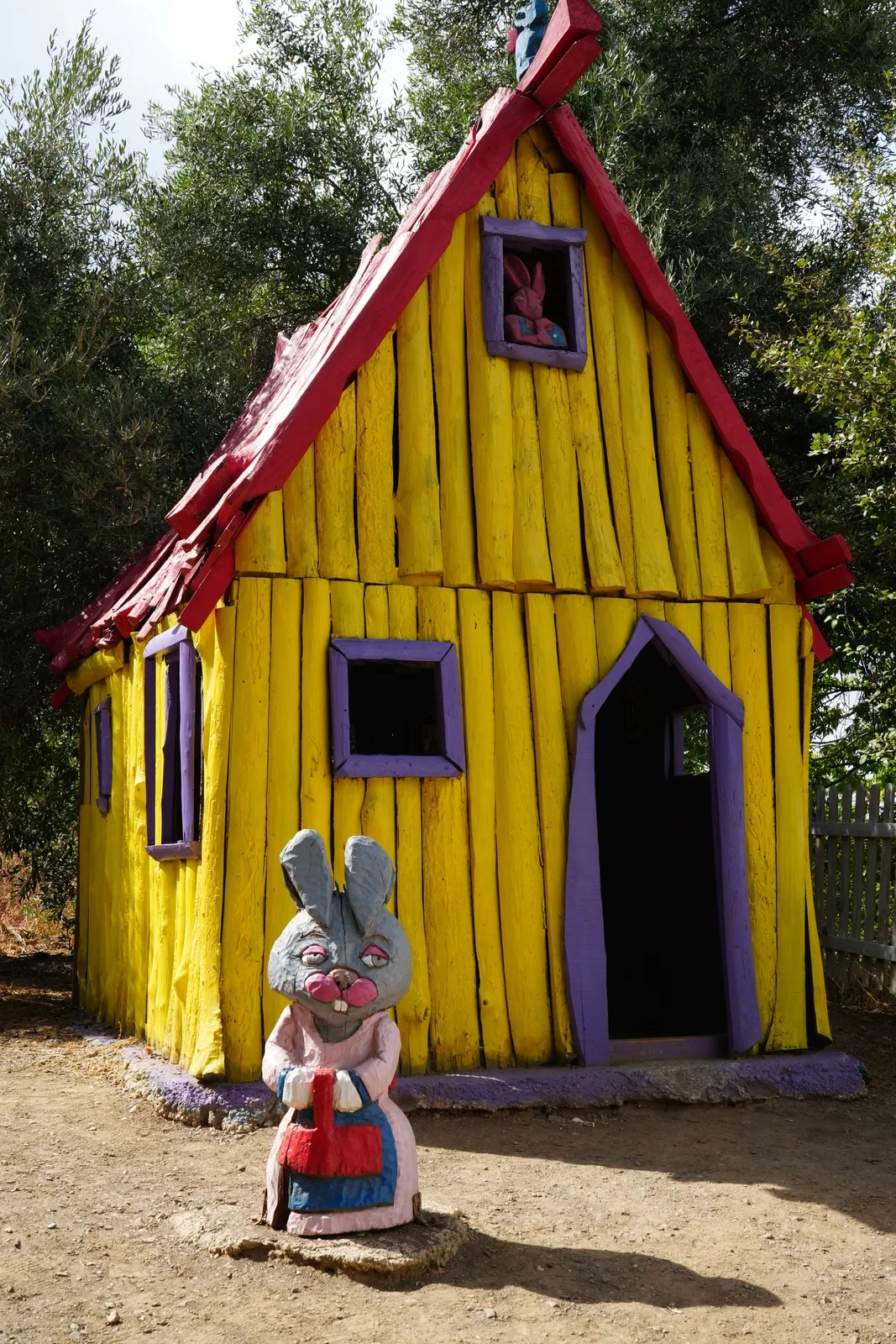 A small yellow wooden house with a red roof and purple door, with a rabbit statue in an apron standing in front