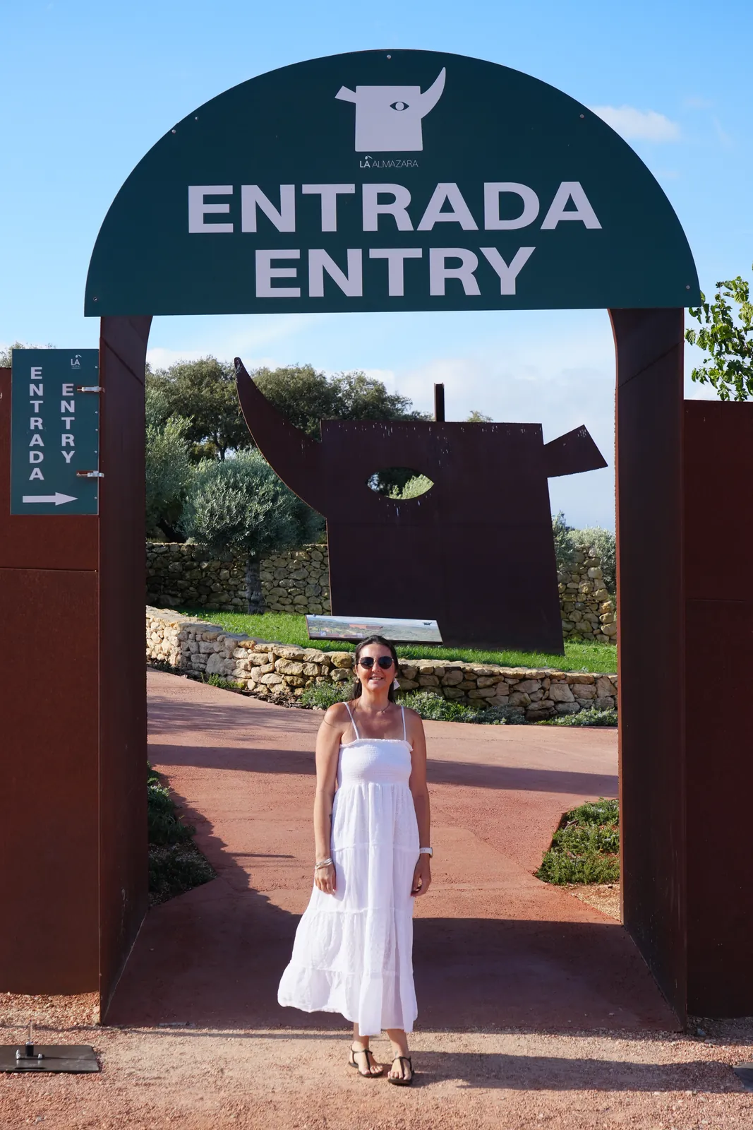 Heidi in a white dress standing under the green ENTRADA ENTRY arch at LA Almazara with a rusted steel bull sculpture behind her