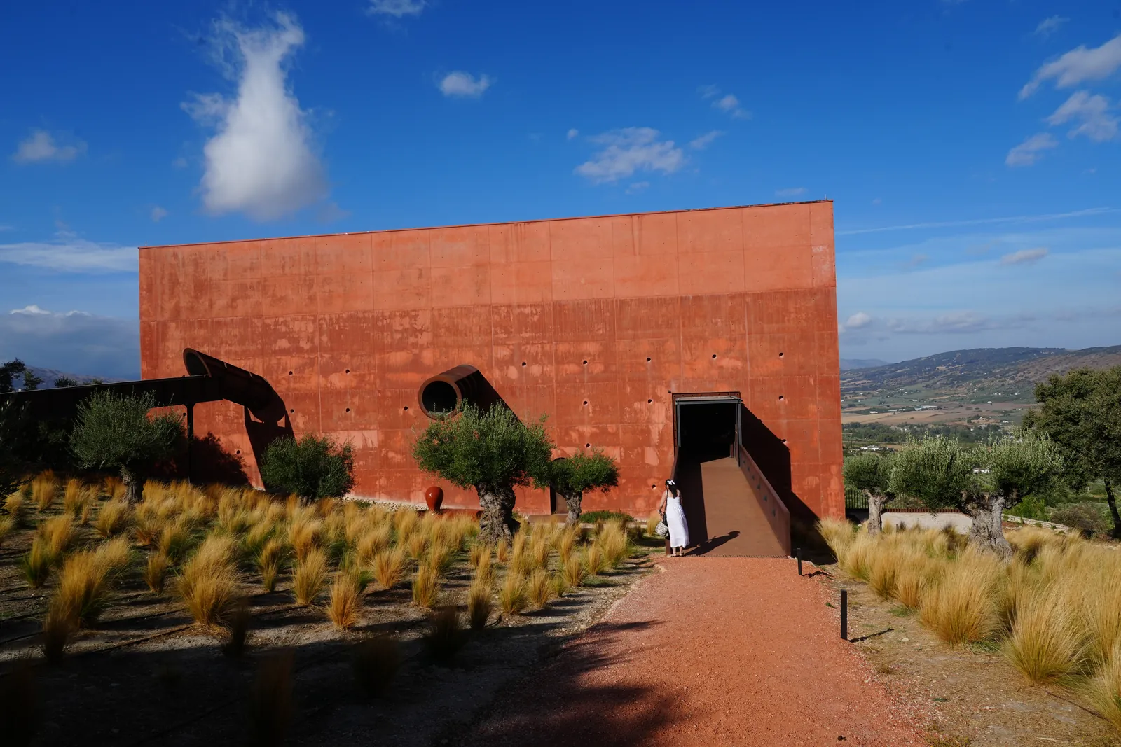 Heidi walking up the steel ramp into the red LA Almazara building, with a pipe sticking out of the wall and olive trees in the grass below