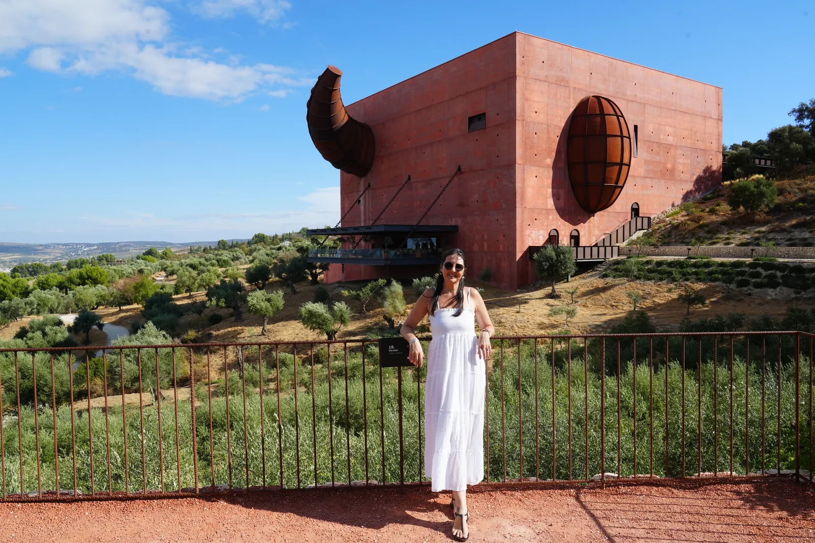 Heidi in a white dress leaning on the rusty mirador railing with the red LA Almazara cube, steel horn and carved window behind her