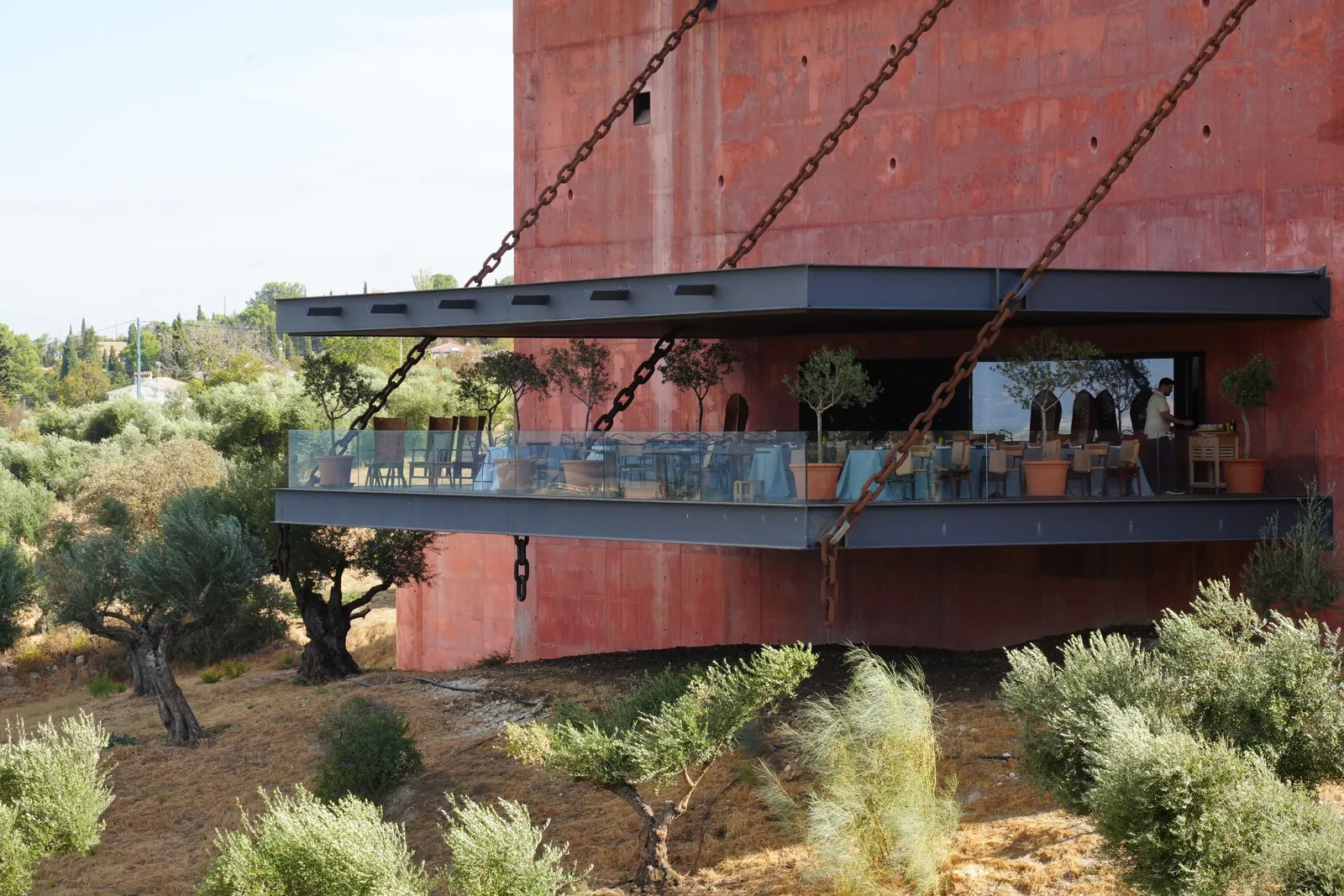 The LA Almazara balcony hanging from long steel chains on the side of the red building, above the olive trees