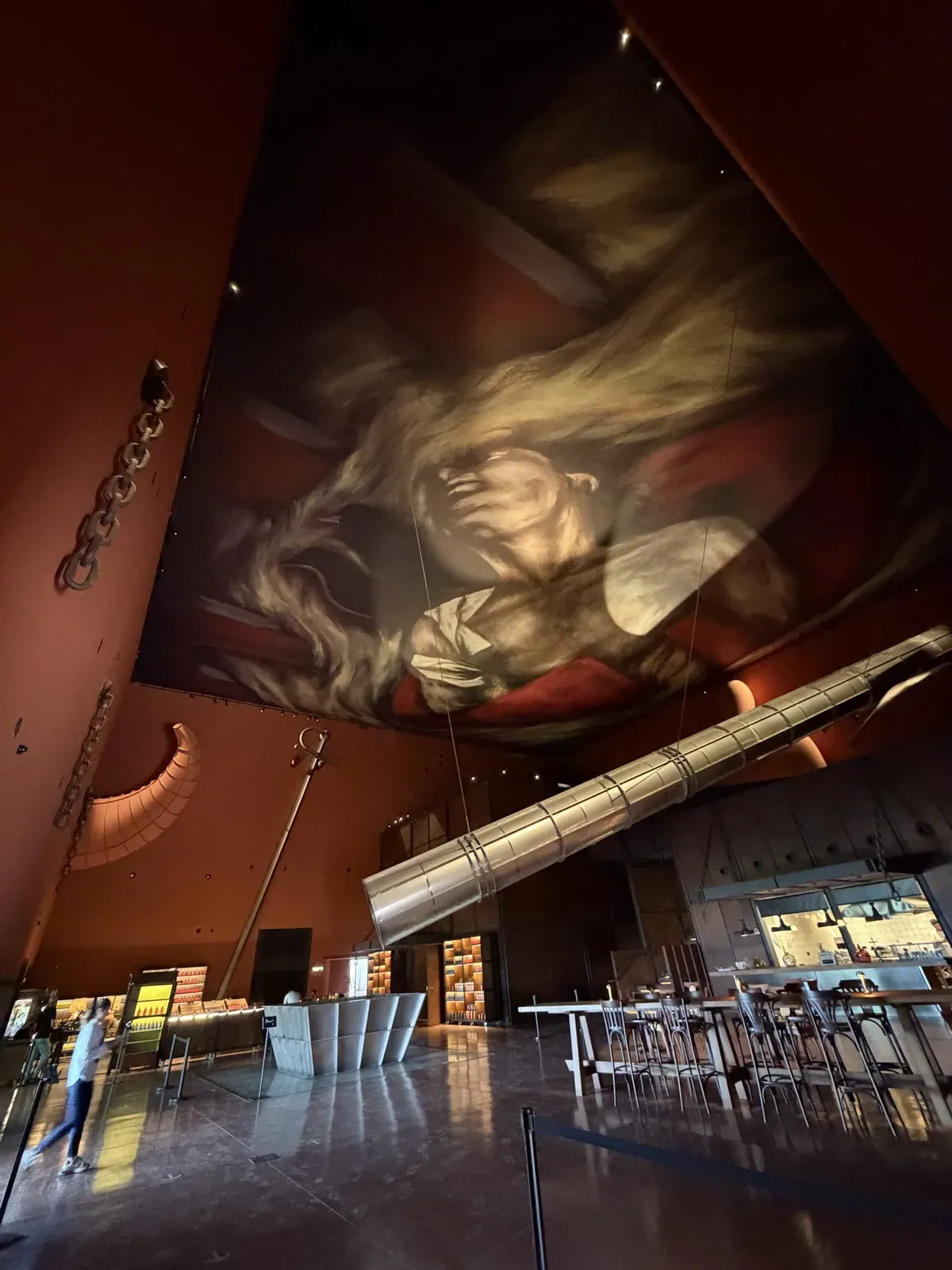 The huge ceiling painting by Ara Starck in the dark main hall of LA Almazara, with a long steel pipe across the hall, a horn on the red wall and the sword in the far left corner