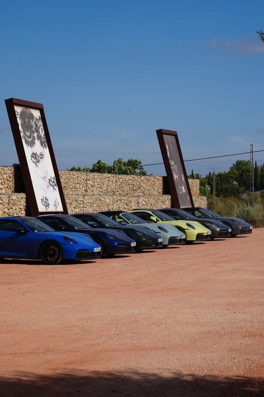 The large red gravel car park at LA Almazara with a row of parked cars in front of two big sun-faded photo panels