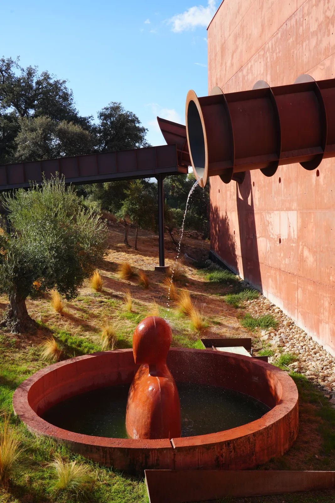 Thin stream of water falling from a steel pipe on the red LA Almazara wall onto a red head-shaped statue standing in a round basin
