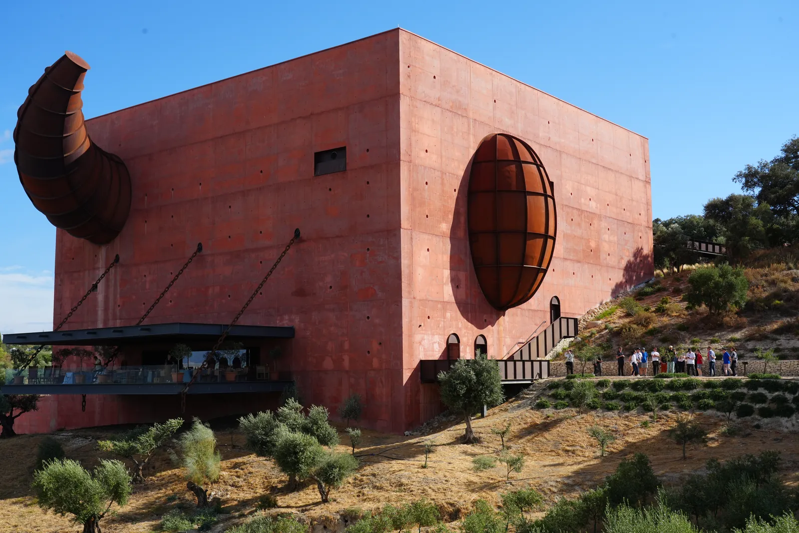 The red LA Almazara cube with its steel bull's horn and oval window above olive groves under a blue sky