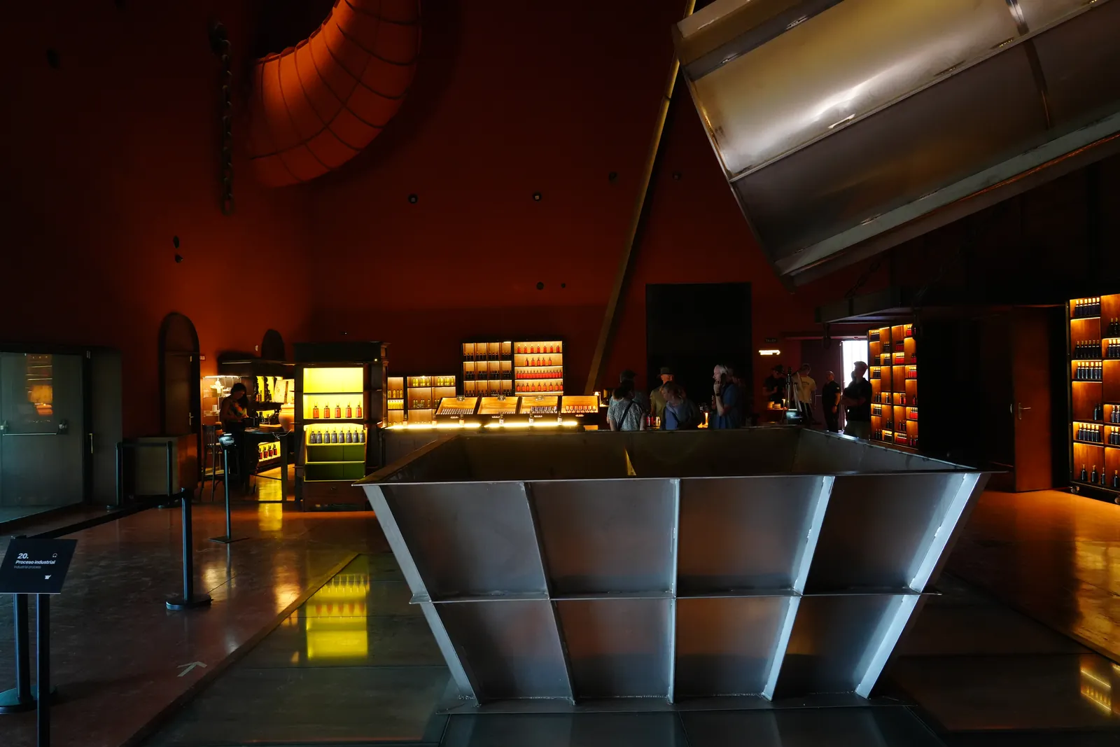 Steel funnel above a glass floor in the dark main hall of LA Almazara, with the bar and shop lit up behind it