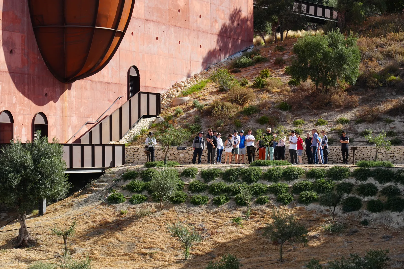 A group of visitors standing on the terrace below the red LA Almazara building with the big oval steel window above them