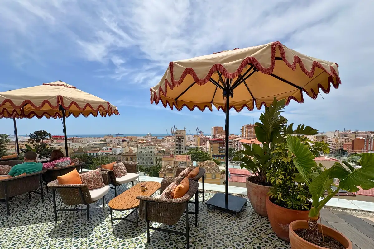 The H10 Croma rooftop terrace in Malaga, with fringed golden umbrellas, wicker seating with orange cushions, Moorish-patterned tiles, and the port and Mediterranean visible beyond