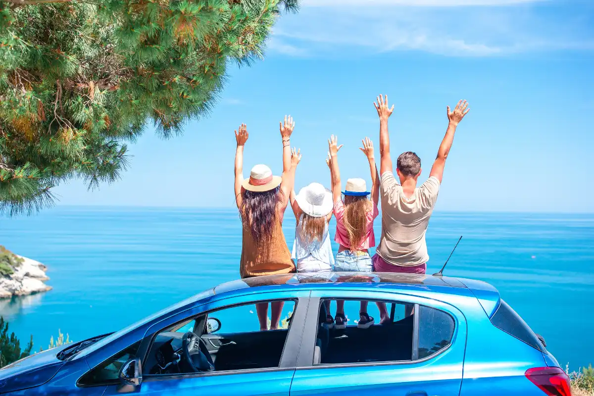 Family of four sitting on top of a car with arms raised, sea view behind them