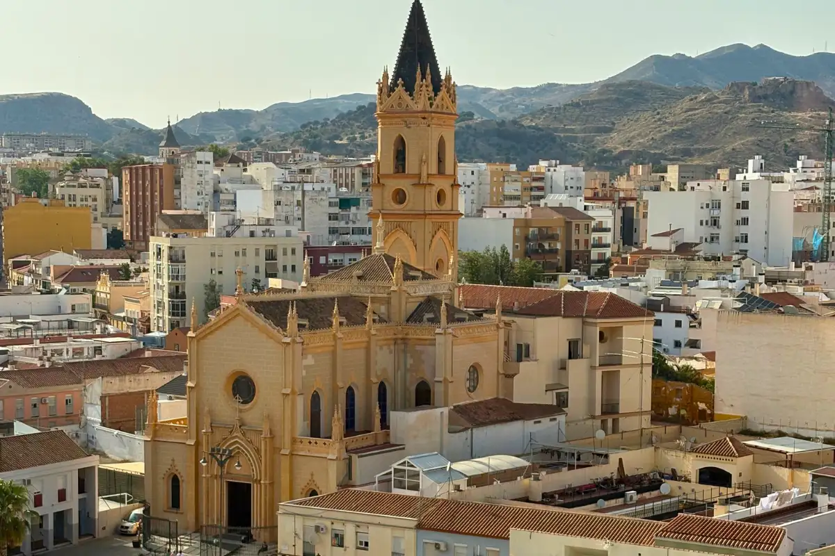 View over the Malaga rooftops from a rooftop terrace, with a golden Gothic church tower in the foreground, the city spreading behind, and Gibralfaro Castle on the hillside in the distance